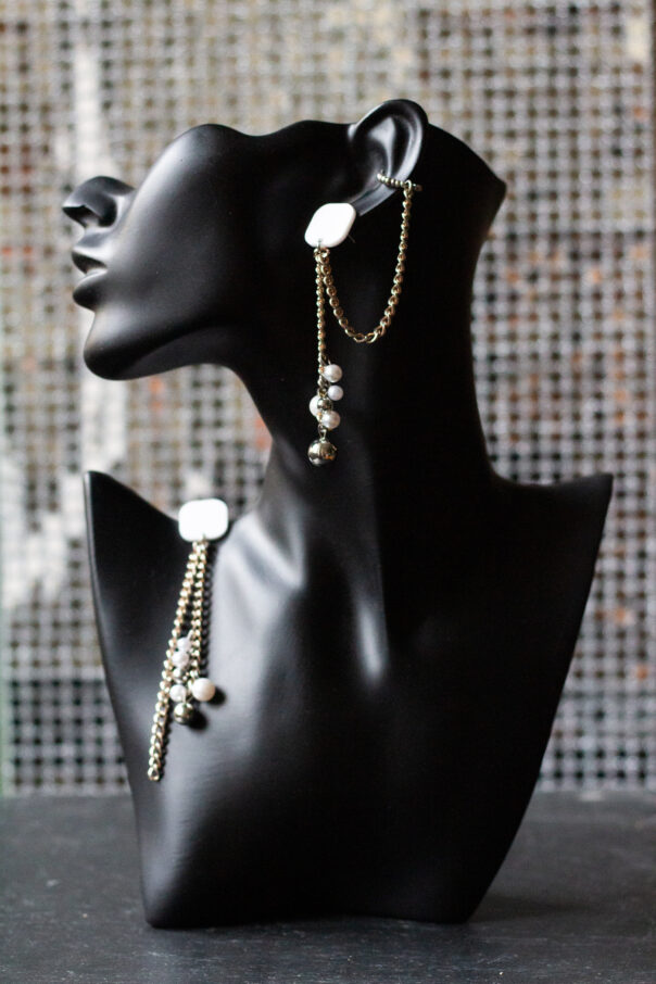 schwarzer Büstenkopf mit Ohrring. Weißer Stecker, Metallketten, unten Perlen aus Kunststoff. Ein Ohrring mit Earcuff