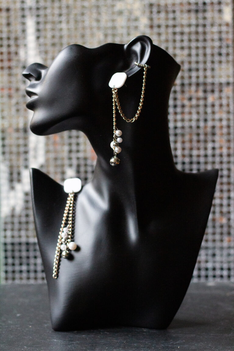 schwarzer Büstenkopf mit Ohrring. Weißer Stecker, Metallketten, unten Perlen aus Kunststoff. Ein Ohrring mit Earcuff